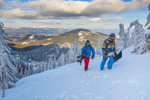 Two happy snowboarders, with backpacks, climb on the mountain slope