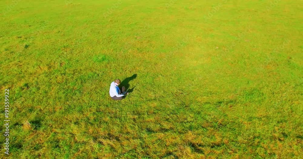 UHD 4k aerial view. Low orbital flight over businessman sitting on green grass with notebook (pad) at yellow rural wheat field. Radial movement.