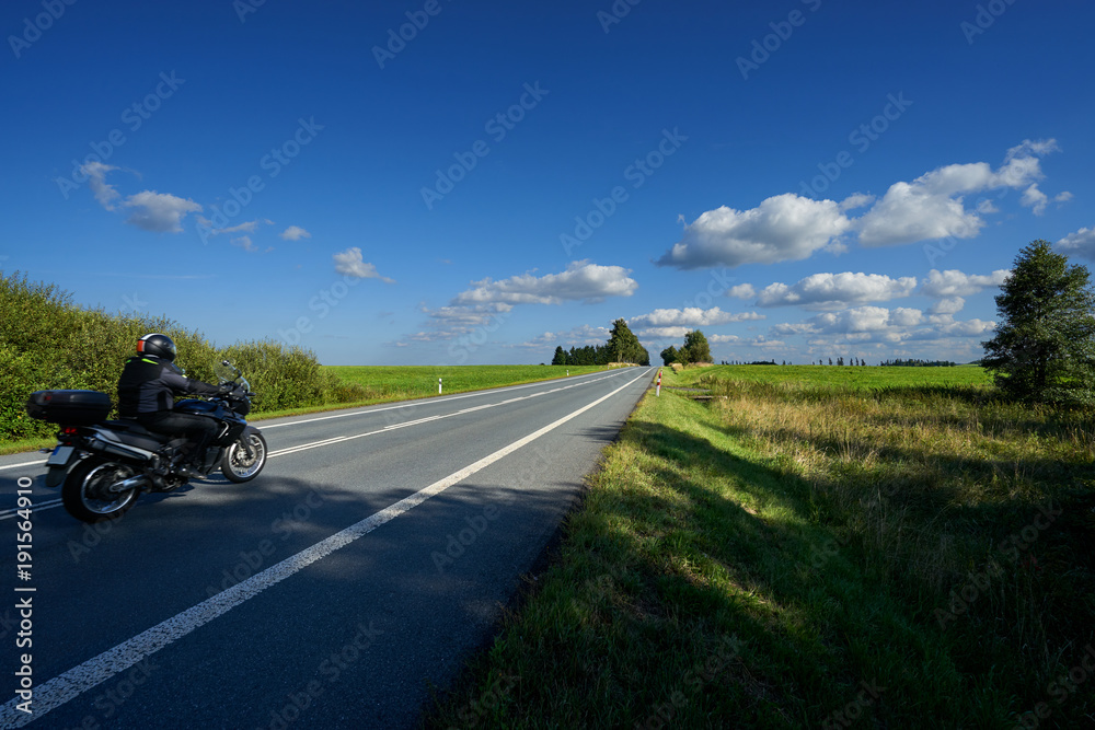 Black motorcycle riding on an empty asphalt road in a landscape on a sunny day with blue sky and white clouds