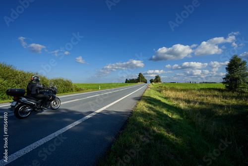 Obraz na płótnie Black motorcycle riding on an empty asphalt road in a landscape on a sunny day with blue sky and white clouds w salonie