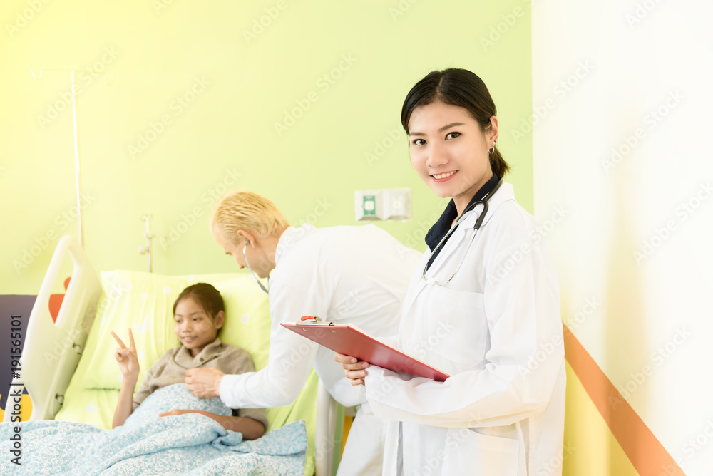 Obraz premium Happy Young asian female doctor standing and holding clipboard,background is caucasian doctor man taking care and examining patient child lying in bed in hospital. Medical and Health care kid concept.