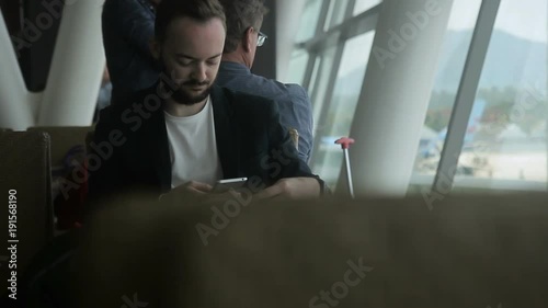 Portrait of male traveller who is writing an email on his smartphone in the airport. Man with beard is wearing white t-shirt and black jacket and is sitting on comfortabe chair near the big window photo