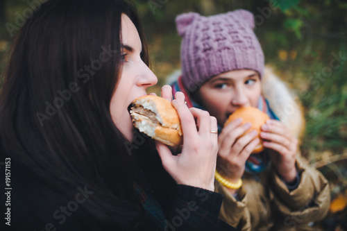 Two sisters eating hamburgers at a picnic in the autumn park.