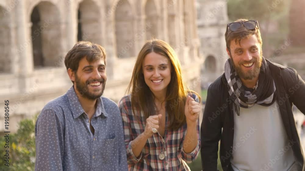 Three young friends tourists jumping posing in front of colosseum in rome at sunset taking selfie pictures photos with dslr camera timer happy beautiful girl long hair slow motion