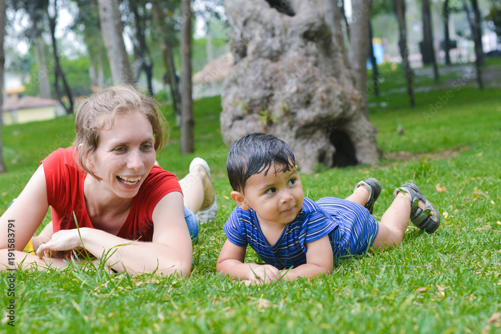 Happy biracial family in the summer park - caucasian mother with her latin son laying on the grass.
