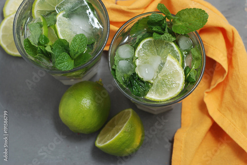 Two Glasses with Mohito and ice with Lime Citrus Fruits. Orange Napkin over Gray Background. Summer cocktail.