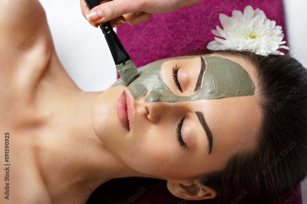 Naklejka premium Woman with clay facial mask in beauty spa. Skincare. Beauty Concept. Close-up portrait of beautiful girl applying facial mask.Facial treatment. Cosmetology. Body care girl's