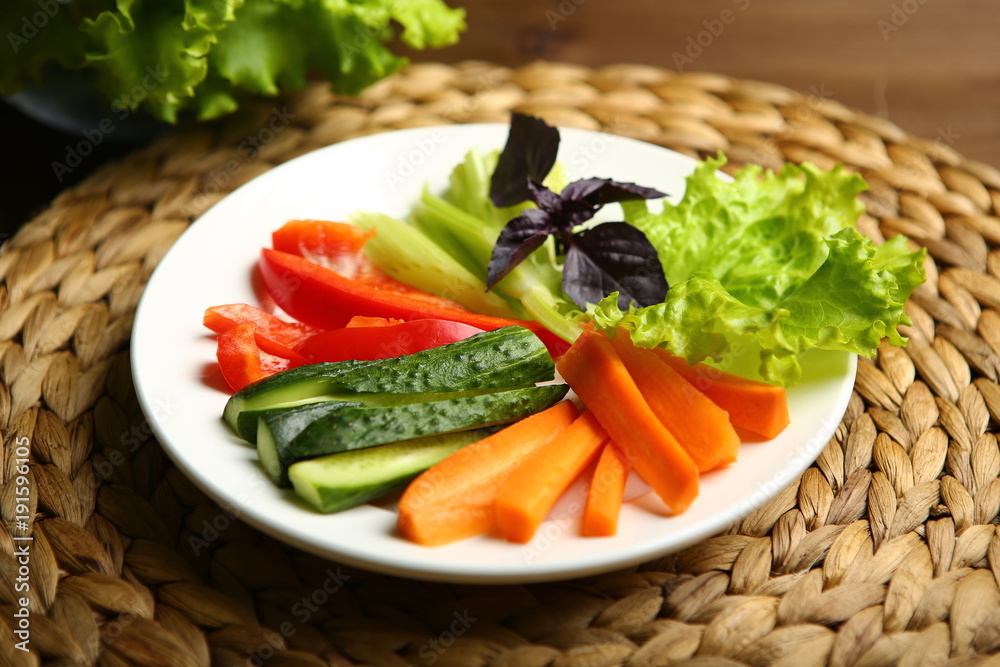 Pepper, carrots, celery, raw cucumber are cut into strips. On the plate. Next to it are lettuce leaves, basil. On a background a wicker stand, a wooden table, a black background. Pepper, carrots, celery, raw cucumber are cut into strips. On the plate. Next to it are lettuce leaves, basil. On a background a wicker stand, a wooden table, a black background.