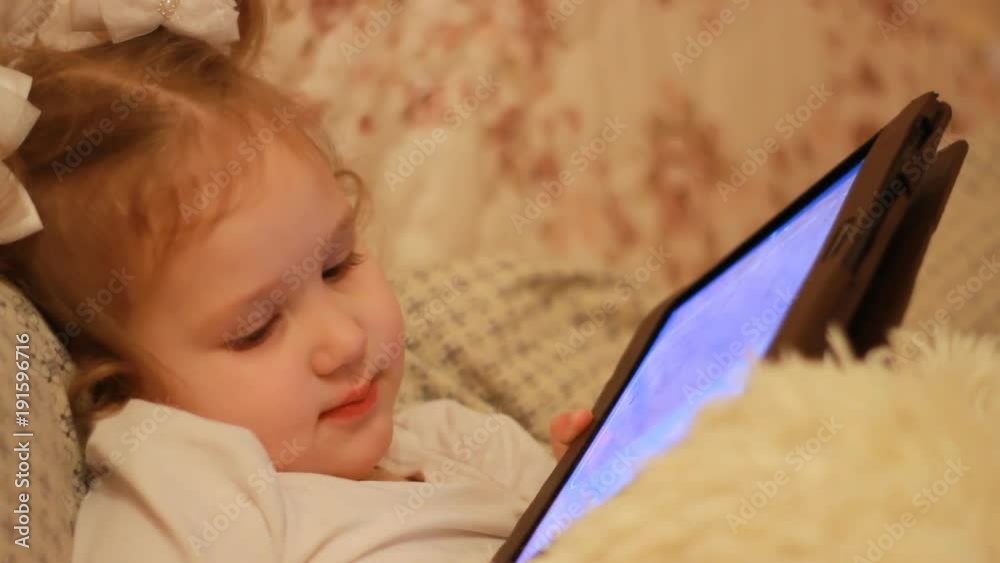 Child Looks cartoons and plays downloaded application on a smart phone close-up. A little cute funny girl lies in bed under the blanket and looks at the white phone screen. Child Looks cartoons and plays downloaded application on a smart phone close-up. A little cute funny girl lies in bed under the blanket and looks at the white phone screen.