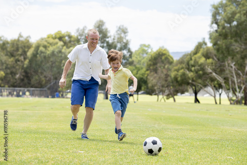 young happy father and excited little 7 or 8 years old son playing together soccer football on city park garden running on grass kicking the ball