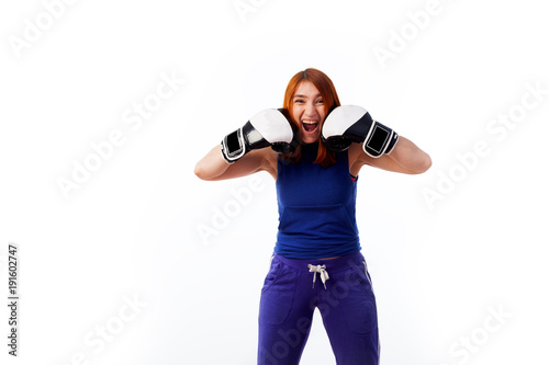 young red-haired woman in sportswear and black and white boxing gloves smiles and weakly strikes herself with two gloves on the head on a white isolated background © Виталий Сова