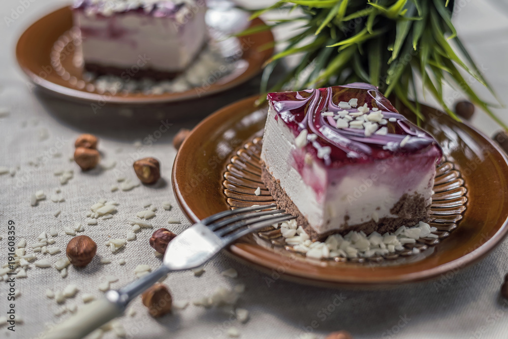 A piece of vanilla cake with chocolate base and cherry glaze decorated with some pieces of white chocolate served in a brown clay dish with silver fork placed on a beige tablecloth. Blurred background A piece of vanilla cake with chocolate base and cherry glaze decorated with some pieces of white chocolate served in a brown clay dish with silver fork placed on a beige tablecloth. Blurred background