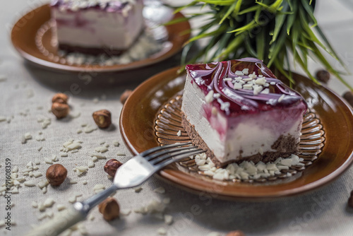 A piece of vanilla cake with chocolate base and cherry glaze decorated with some pieces of white chocolate served in a brown clay dish with silver fork placed on a beige tablecloth. Blurred background