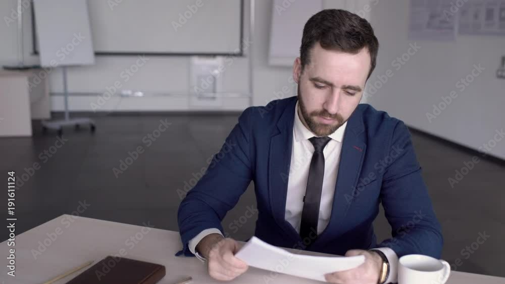 Portrait of businessman is reading a report in conference room in the modern office. Professional in blue suit with tie is sitting at the wooden table. holding the papers in his hands and looking to Portrait of businessman is reading a report in conference room in the modern office. Professional in blue suit with tie is sitting at the wooden table. holding the papers in his hands and looking to
