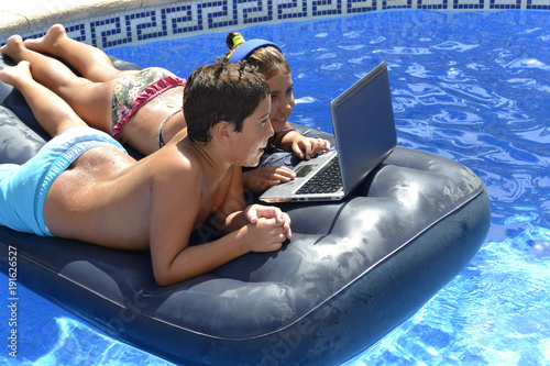 young boy and girl watching a movie on a partitive computer lap top  swimming in the pool on an inflatable mattress photo