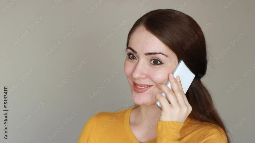 Beautiful young brunette woman talks on the mobile phone and smiling. Happy girl with a white smartphone in her hand on a light background Beautiful young brunette woman talks on the mobile phone and smiling. Happy girl with a white smartphone in her hand on a light background