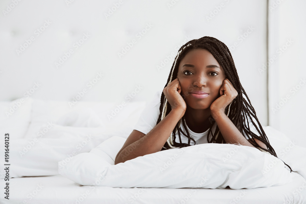 A beautiful African woman lies on her stomach, on a bed, in a white bedroom. Good morning, vacation at home. The girl is wearing a T-shirt, pigtails on her head. Healthy sleep. Portrait. A beautiful African woman lies on her stomach, on a bed, in a white bedroom. Good morning, vacation at home. The girl is wearing a T-shirt, pigtails on her head. Healthy sleep. Portrait.