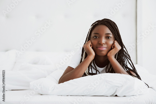 A beautiful African woman lies on her stomach, on a bed, in a white bedroom. Good morning, vacation at home. The girl is wearing a T-shirt, pigtails on her head. Healthy sleep. Portrait.