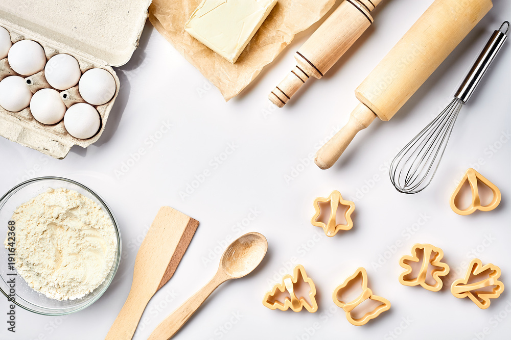 Bowl with wheat flour, rolling pin, whisk, eggs, butter, cookie cutters. Top view on a white table with a copy space Bowl with wheat flour, rolling pin, whisk, eggs, butter, cookie cutters. Top view on a white table with a copy space