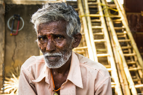Portrait of Indian elder man with traditional bindi as a third eye, white beard, bamboo ladders on the background and a saw hanging from the wall in Mysore, Karnataka, India. Rural village hard worker