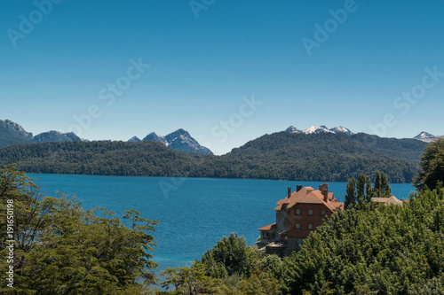 Brick house facing a blue lake, in wooded area and snowy mountains in the background