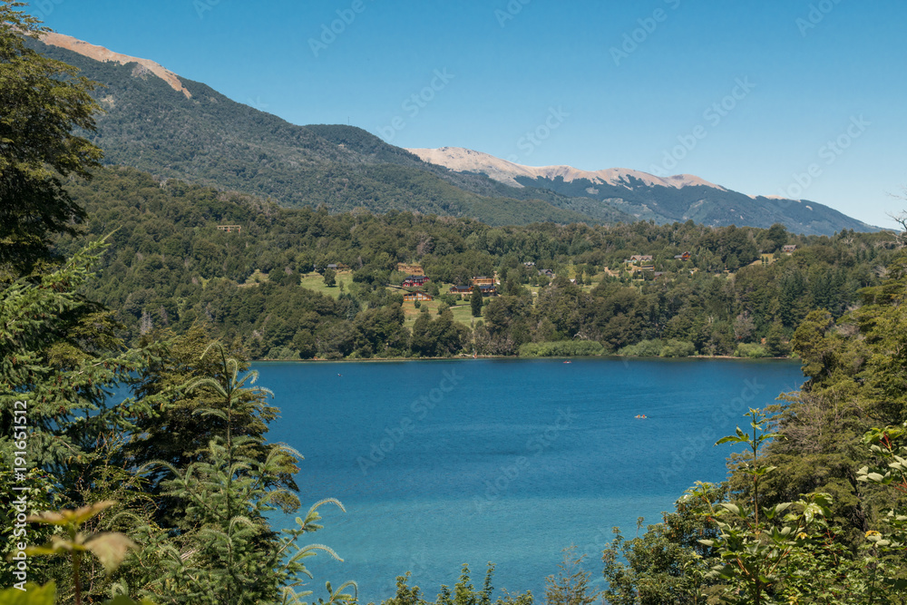 Landscape of a turquoise lake with houses in the background, trees and mountains
