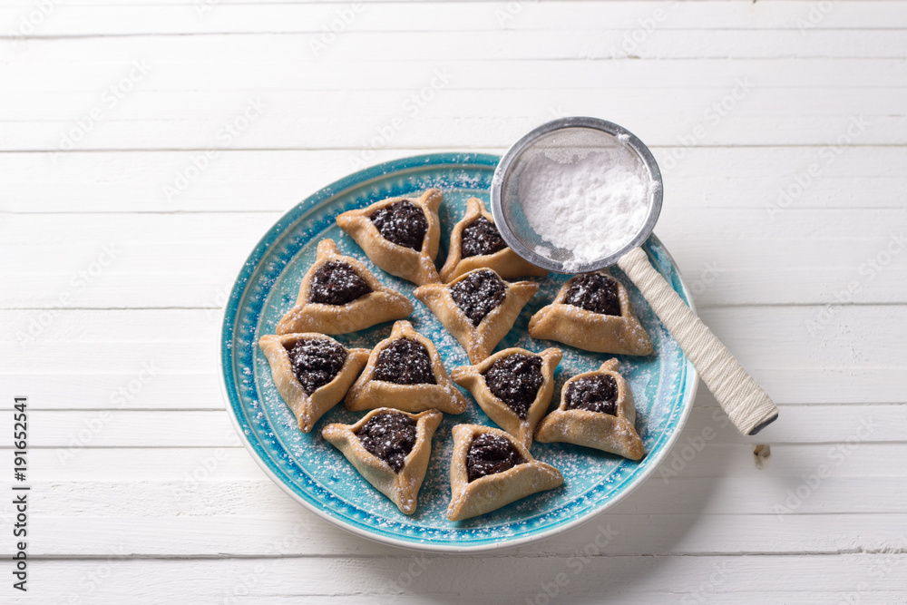 Traditional sweets for the Jewish holiday Purim. Hamantashen cookies or ears of Haman, triangular cookies with poppy seeds and raisins on a blue plate, top view, free space