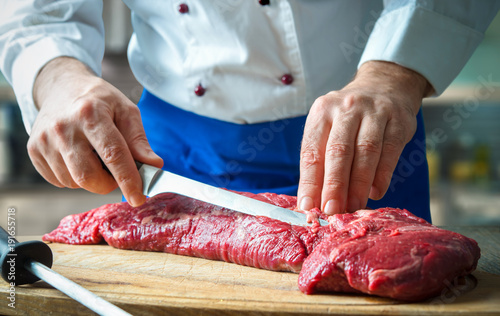 Hands of male chef in uniform cutting big piece of beef