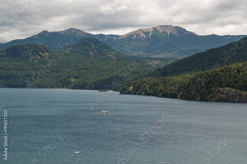 Lake landscape on cloudy day, with a white catamaran and forested mountains in background