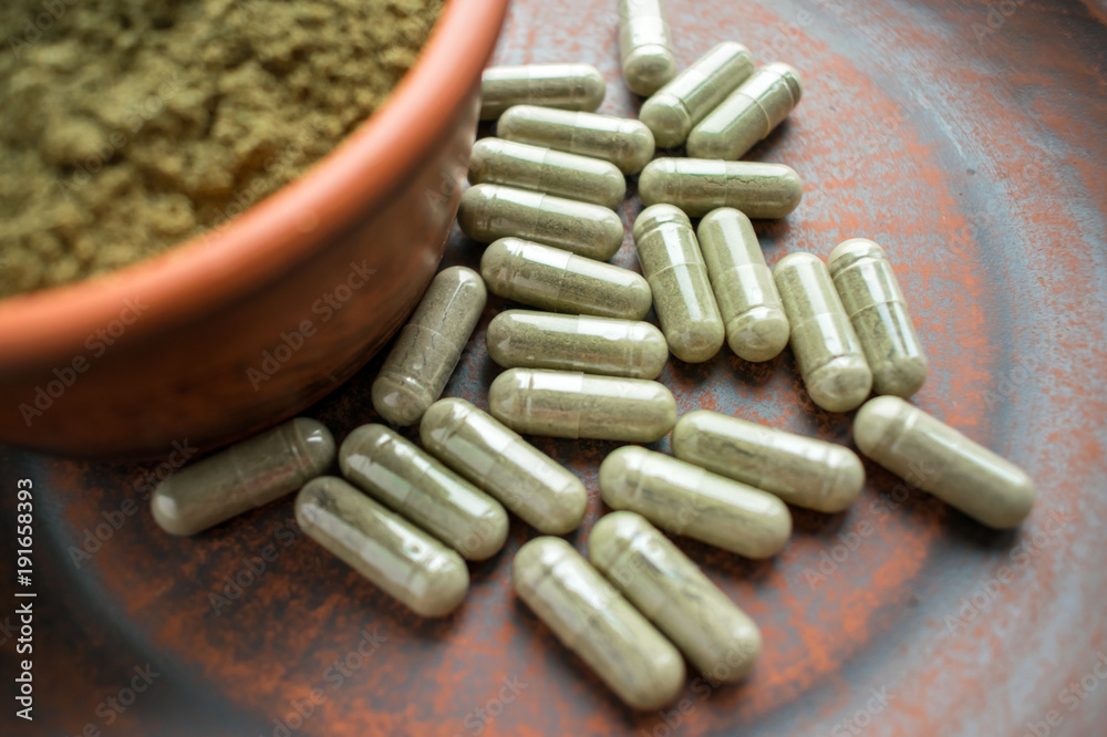 Supplement kratom green capsules and powder on brown plate. Herbal product alt-medicine kratom is opioid. Home alternative pain remedy, opioid addiction, painkiller, overdose. Selective focus