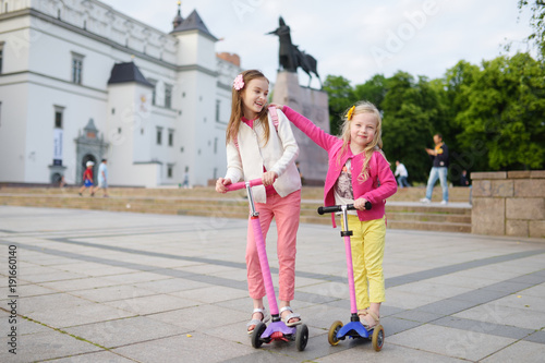 Small children learning to ride scooters in a city park on sunny summer evening. Cute little girls riding rollers.