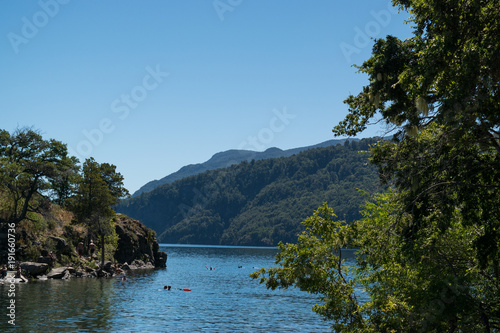 Landscape of a lake with an island, people swimming and mountains in the background