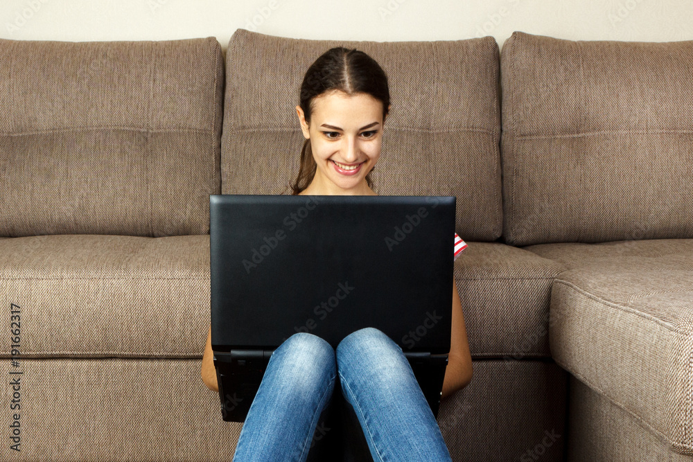 Beautiful girl in casual clothes is using a laptop and smiling while sitting on the floor near the couch at home Beautiful girl in casual clothes is using a laptop and smiling while sitting on the floor near the couch at home