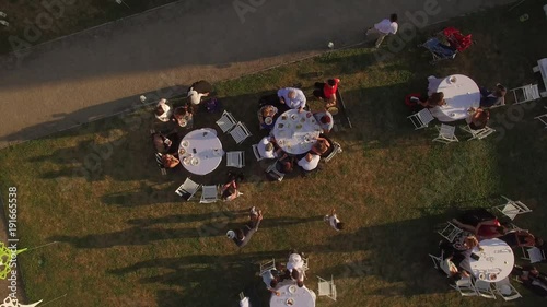 Low flight shot over people sitting at tables moving on green lawn at open air bbq picnic party in outside yard restaurant or cafe top sun view. Local food and wine tasting feast festival in Europe