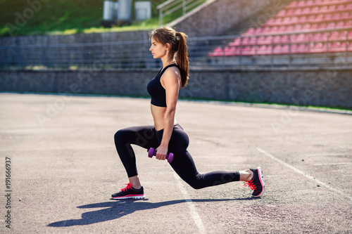Fitness girl with ultra violet dumbbells doing warm up on the sadium before training, stretching body muscles. Female athlete preparing legs for cardio workout. Active girl doing exercises outdoor.