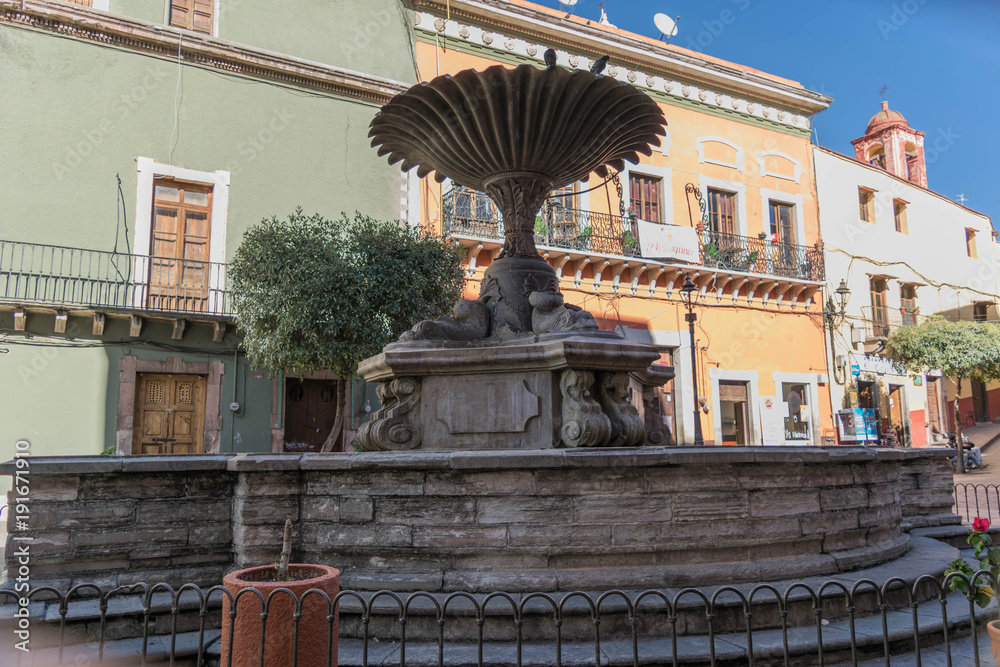 Fototapeta premium The stone fountain in the Plaza del Baratillo, on a sunny day, with colorful buildings in the background, in Guanajuato, Mexico