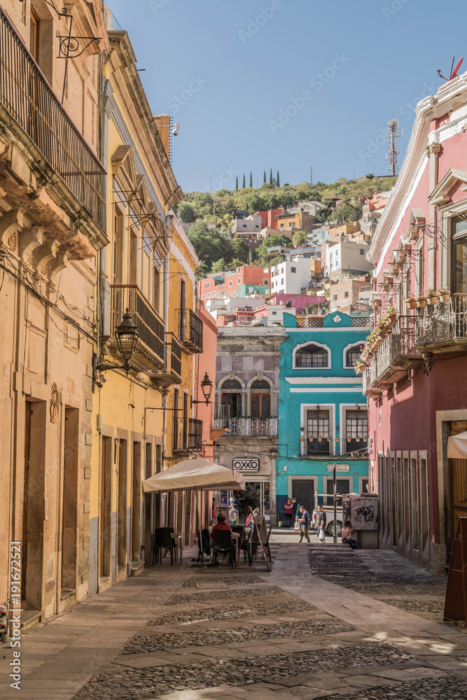 A charming cobblestone pedestrian street with colorful buildings, outdoor table, and a hill dotted with colorful homes, in the background, in Guanajuato, Mexico