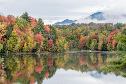 Vibrant fall colored forest with dark mountain behind it. Both mountain and trees full of fall foliage reflecting in a smooth lake in New England.