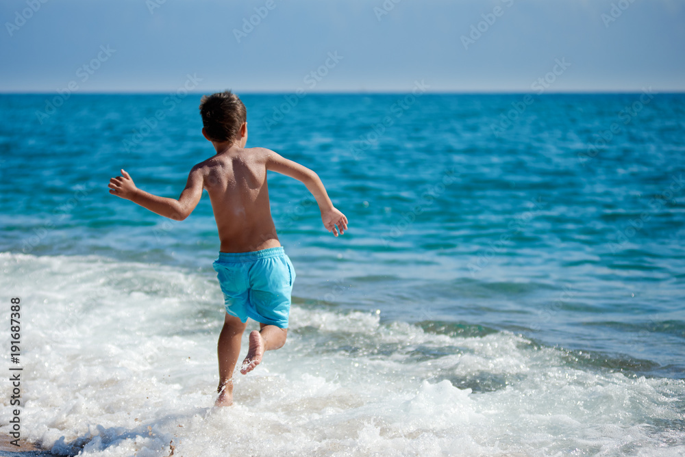 Caucasian boy is running in the water along the sea shore. Back view.