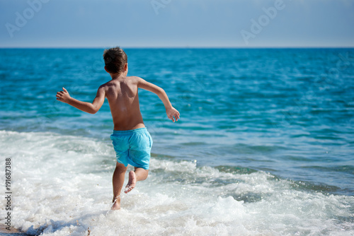 Obraz na płótnie Caucasian boy is running in the water along the sea shore. Back view. w salonie