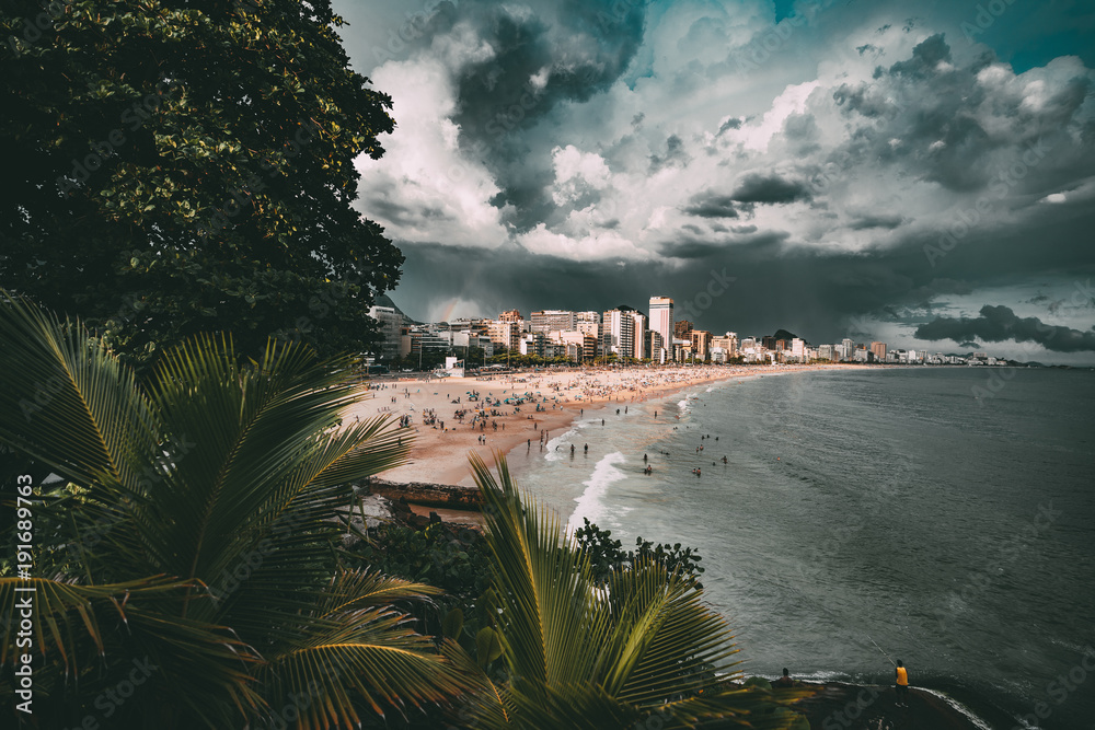 Wide-angle shooting of stunning cityscape during the storm in Rio de Janeiro: coastline with Leblon and Ipanema beaches, swimming and tanning people, dramatic sky with heavy rain in distance, palms