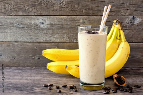 Coffee, banana smoothie in a tall glass with coffee beans and bananas in background. Side view, against a rustic wood background.