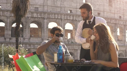 Happy young couple tourists tasting coffee chatting with funny italian elegant waiter sitting at bar restaurant in front of colosseum in rome at sunset joking and laughing having fun