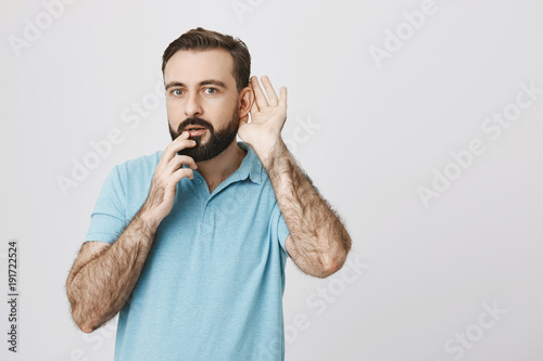 Curious bearded guy holding hand near ear to hear rumor better with fingers on lips, expressing excitement of what he just overheard, over gray background. Man stands behind door and eavesdropping.
