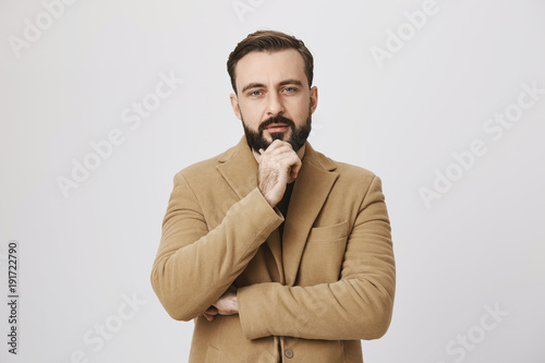 Bearded man in coat looking interested in something holding his hand on chin looking at camera over white background. Middle aged person thinking about investitions he recently made. © Cookie Studio