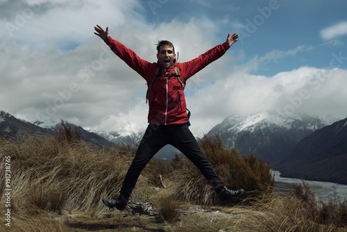 Hombre joven con chaquetón rojo saltando frente a montañas con picos nevados en Nueva Zelanda