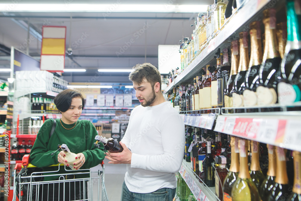 A young beautiful couple buys alcohol in the store. A couple chooses wine at a supermarket.