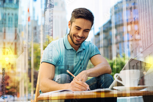 Young writer. Cheerful smart creative young author feeling excited and glad while sitting alone in a quiet place and smiling while writing the last chapter of his future book