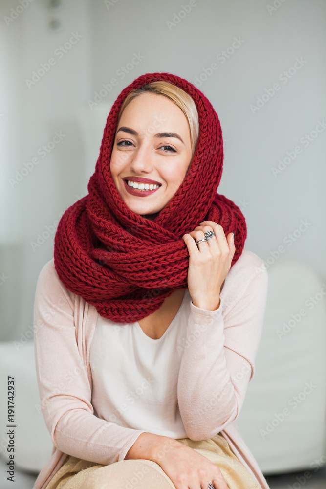 Young woman in warm hand knitted snood scarf at home. Beautiful girl knitting in cozy flat wearing casual clothes, big deep red scarf. Hygge, comfort and home concept. Young woman in warm hand knitted snood scarf at home. Beautiful girl knitting in cozy flat wearing casual clothes, big deep red scarf. Hygge, comfort and home concept.