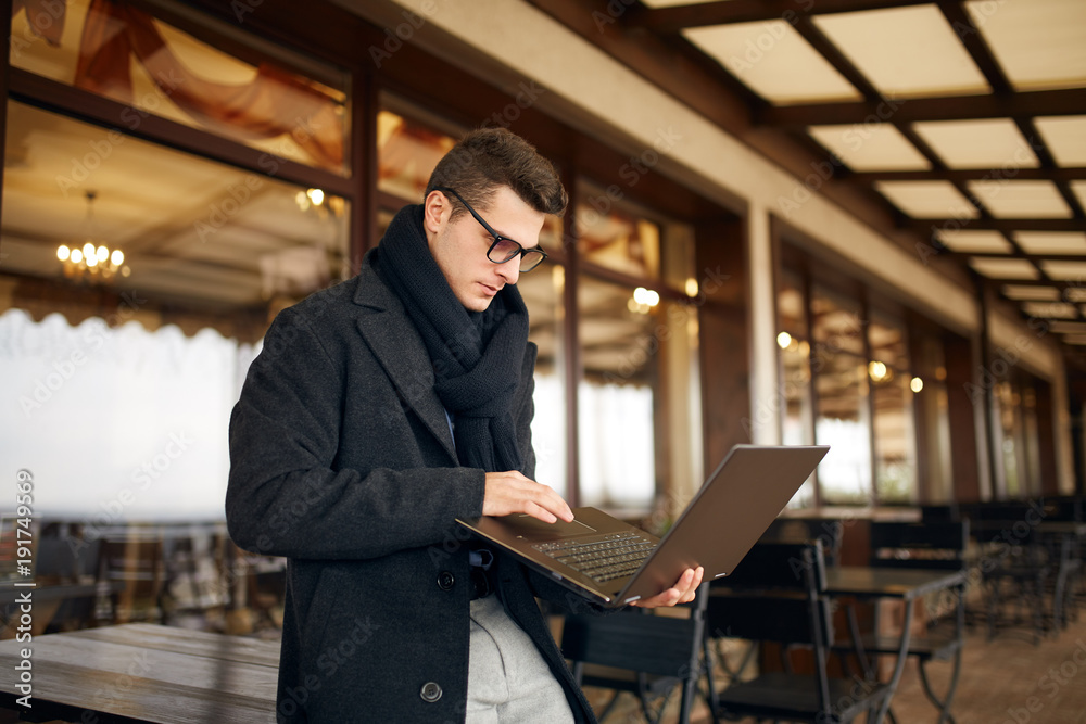 Handsome attractive businessman using a laptop on office terrace. Stylish man in glasses coat and scarf standing on balcony with laptop. Smart casual clothes. Business theme. Handsome attractive businessman using a laptop on office terrace. Stylish man in glasses coat and scarf standing on balcony with laptop. Smart casual clothes. Business theme.